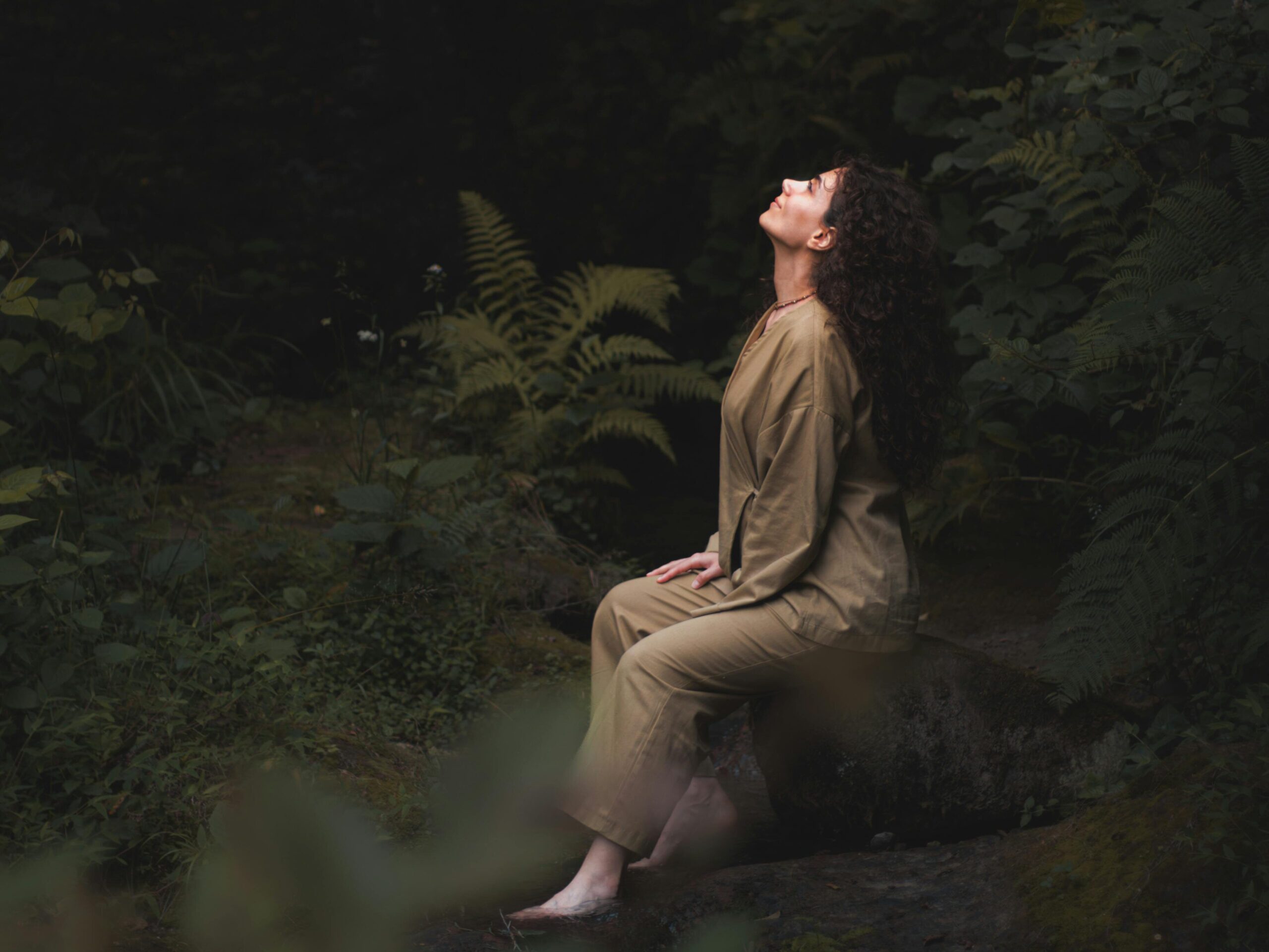 A woman sitting in a lush green forest, looking up at the canopy.